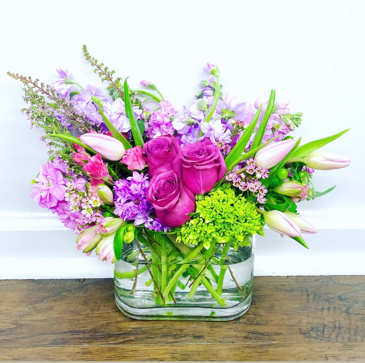 Bouquet of purple and green flowers in a clear vase on a wooden surface with a white background