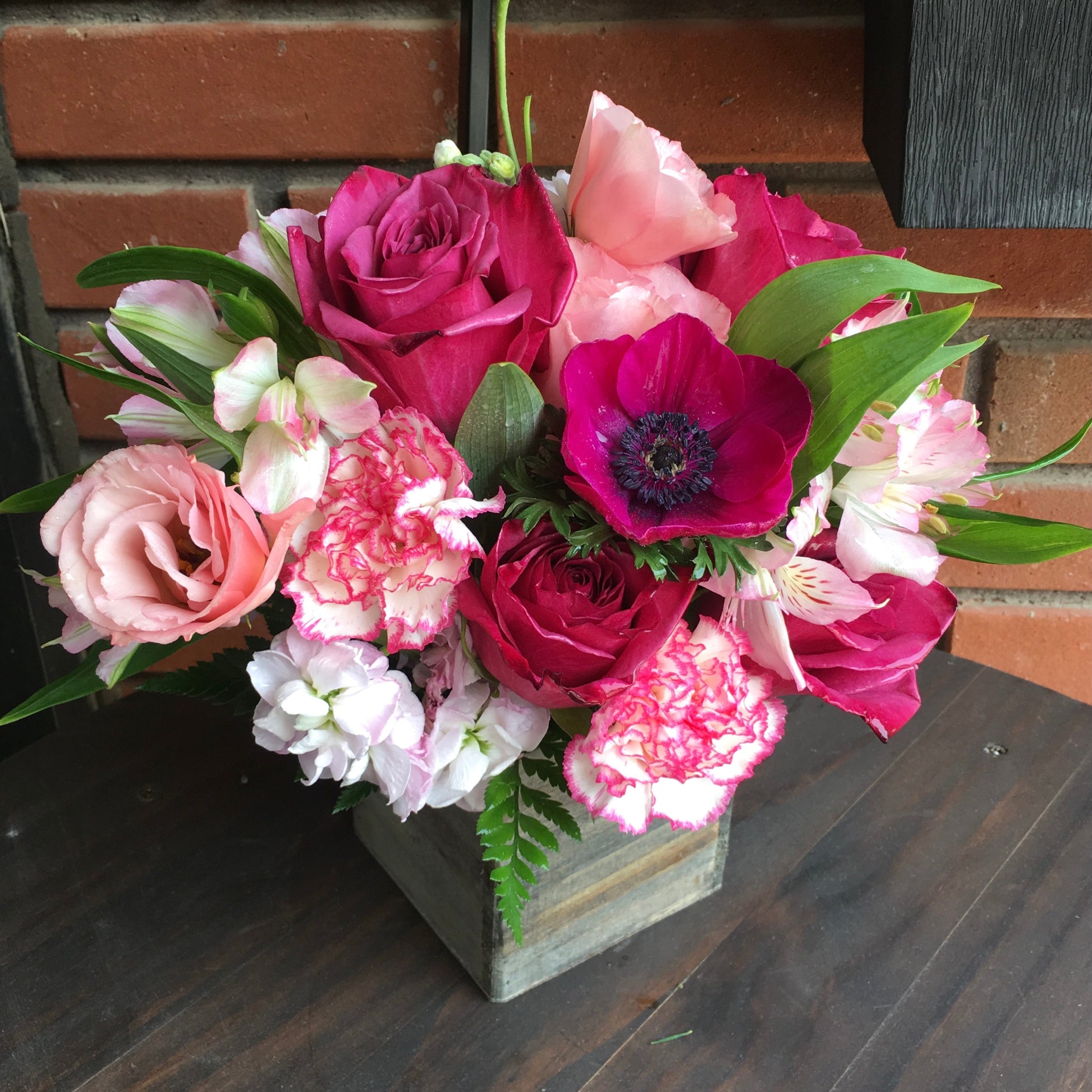 Colorful flower arrangement in a wooden box on a wooden surface with a brick wall background.