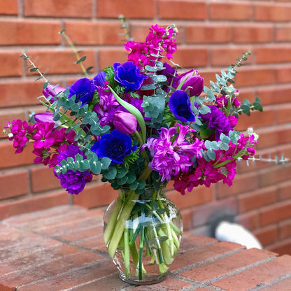 Bouquet of purple and blue flowers in a clear vase against a brick wall from Miami Beach florist for flower delivery
