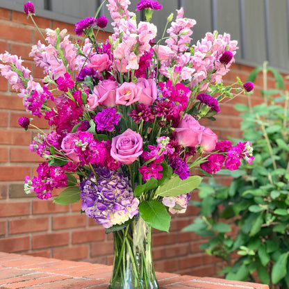 Bouquet of pink and purple flowers in a clear vase on a brick surface with a brick wall and greenery in the background for flower delivery from a Miami Florist