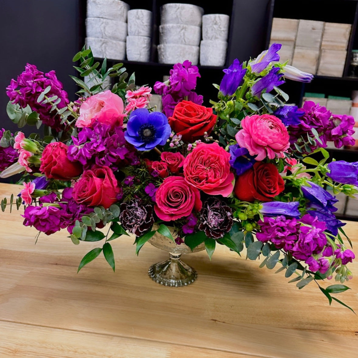 Colorful flower arrangement in a silver vase on a wooden surface with a store shelf in the background from the best miami flower shop