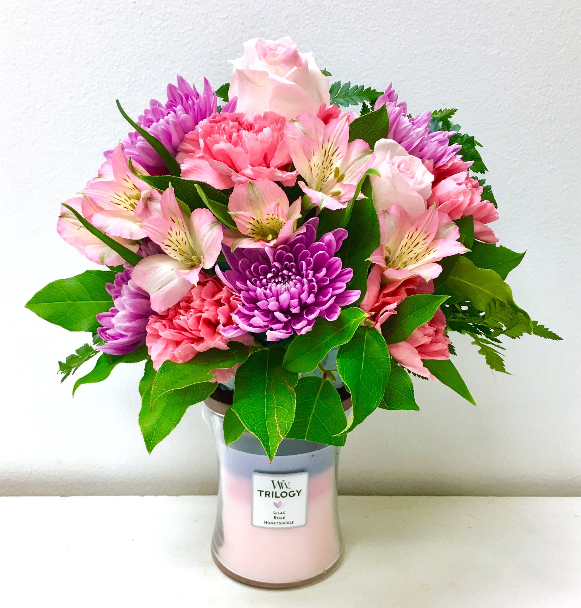 Bouquet of pink and purple flowers in a Trilogy candle container on a white background from Miami florist