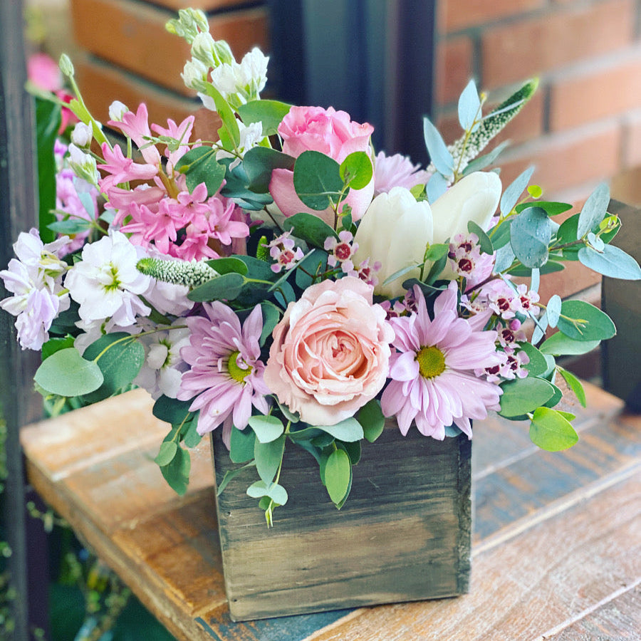 Floral arrangement in a wooden box on a table with a brick wall background to send flowers in Sweetwater Florida