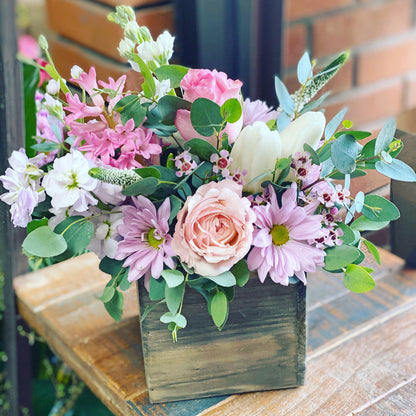 Floral arrangement in a wooden box on a table with a brick wall background to send flowers in Sweetwater Florida